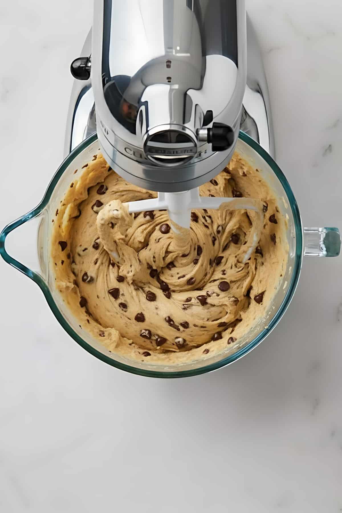 Creamy chocolate chip cookie dough being mixed in a glass bowl with an electric hand mixer.