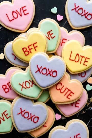 Delicious heart-shaped cookies decorated with pastel icing and sweet words like "LOVE" and "XOXO" on black marble background.